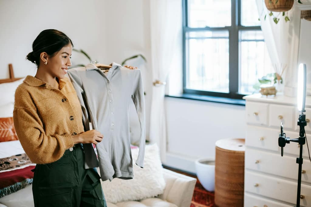 Positive ethnic female blogger demonstrating jacket on hanger during live stream on mobile phone placed on shining lamp